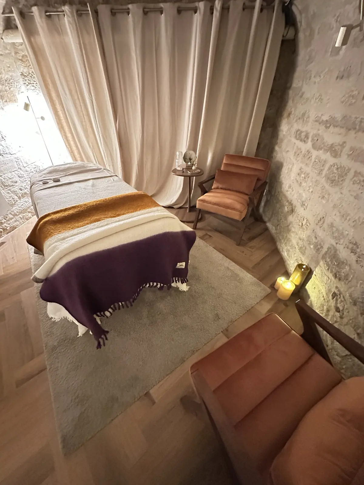 Cabine de l'atelier Pimp My Derm du passage Choiseul à Paris : table de massage, murs en pierre, rideaux en lin, fauteuils terracotta et bougies
